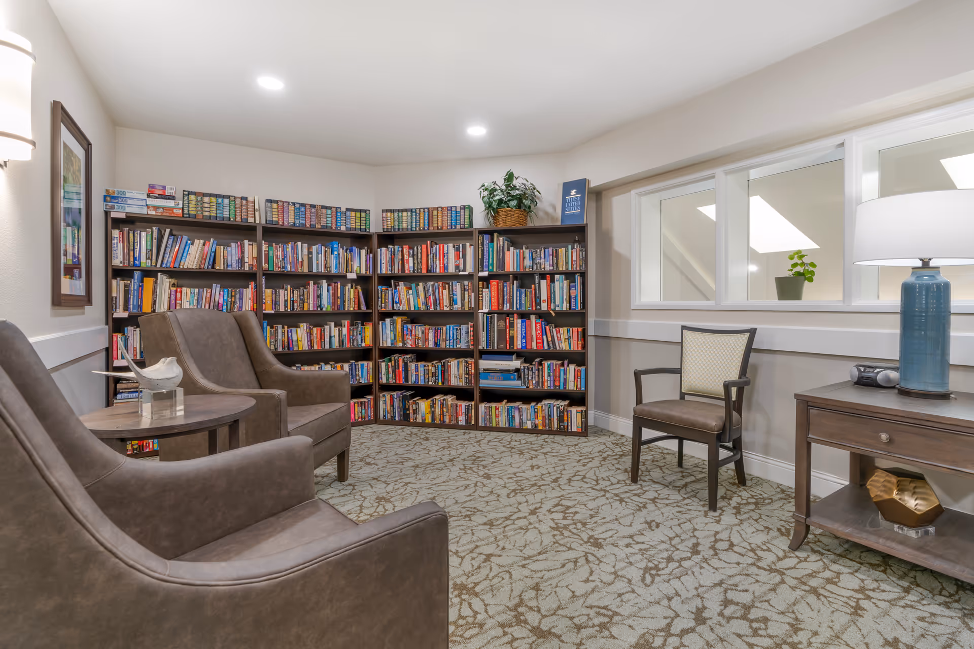 Cozy library-style sitting room with bookshelves along the back wall, upholstered chairs, a side table and a lamp.