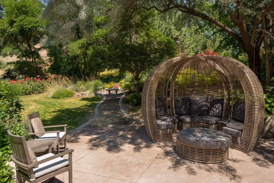 Outdoor patio area with two wooden chairs with cushions on the left and a round wicker seating pod with cushions on the right, surrounded by green trees, bushes, and a garden path in the background.