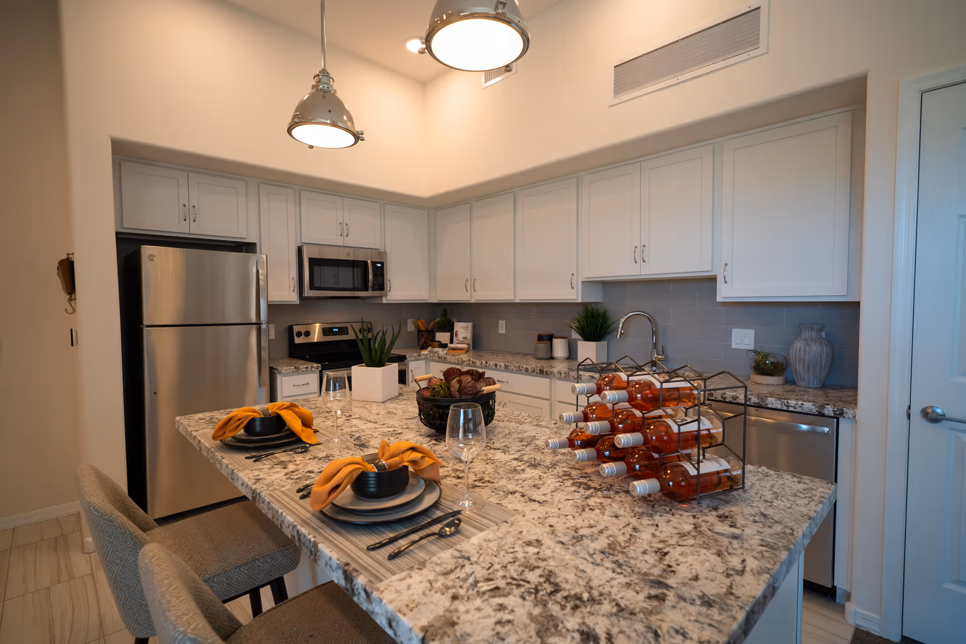 Modern kitchen with white cabinets, stainless steel refrigerator, microwave, and dishwasher. A large granite island countertop with two place settings featuring black bowls, plates, silverware, and orange napkins. The island also has a wine rack with multiple bottles of rosé wine and decorative plants. Two pendant lights hang above the island.