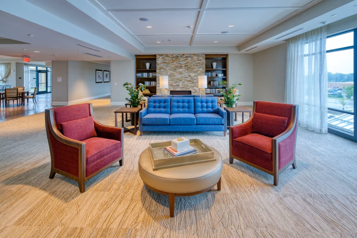 A bright and spacious living room area featuring a blue tufted sofa flanked by two red armchairs. In front of the seating is a round beige ottoman with a tray holding books and a decorative item. Behind the sofa is a stone accent wall with built-in shelves filled with books and decorative vases, and two table lamps. Large windows with sheer curtains allow natural light to fill the room, and there are plants on either side of the sofa. The room has a beige carpet and light-colored walls.