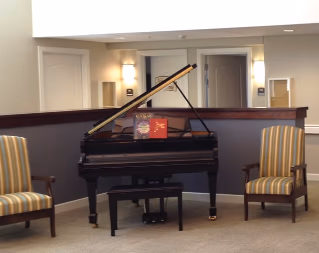A black grand piano with its lid open is centered in a room, flanked by two striped upholstered armchairs. The background shows a half wall with a wooden ledge and two doorways with wall sconces on either side.
