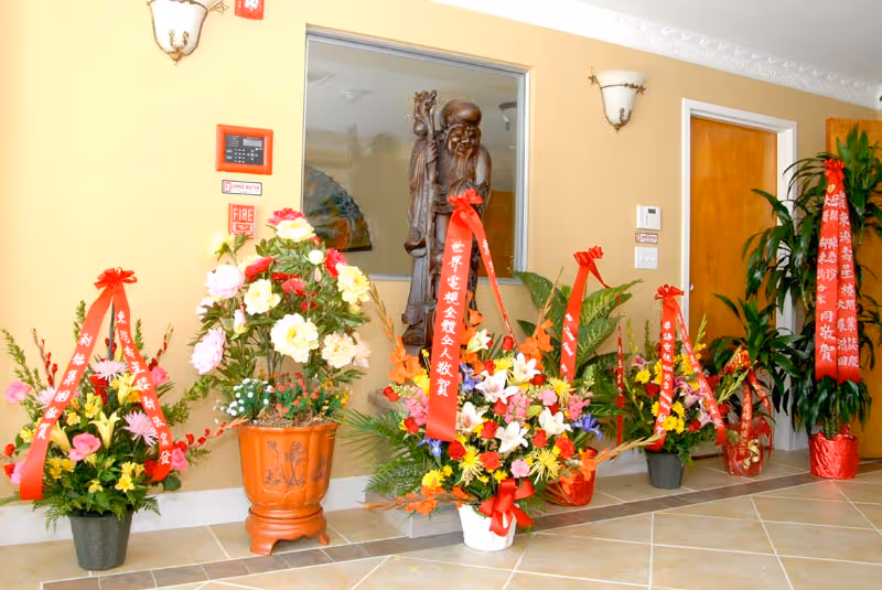 An interior hallway with beige walls and tiled floor featuring several large floral arrangements with red ribbons containing Chinese characters. A wooden statue is visible in a recessed niche in the wall, and there are two wall-mounted light fixtures above the flowers. A wooden door and some green plants are also present.