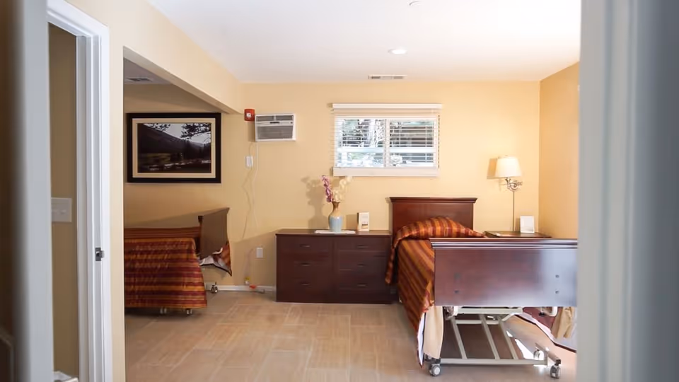 A room with two hospital-style beds with striped bedspreads, a wooden dresser with a vase of flowers, a window with blinds, a wall-mounted air conditioning unit, a floor lamp, and a framed picture on the wall.