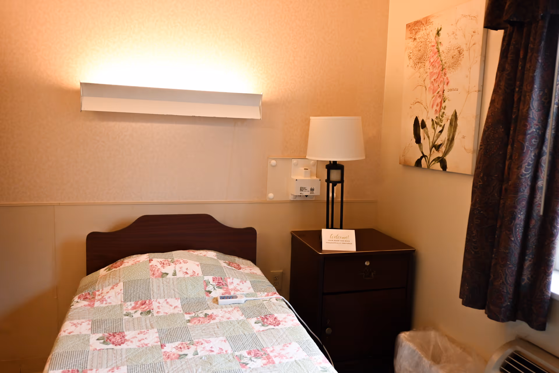 A small bedroom with a single bed covered in a floral patchwork quilt. Next to the bed is a dark wooden nightstand with a lamp and a small sign on it. A floral painting hangs on the wall above the nightstand, and a window with dark patterned curtains is partially visible on the right side.