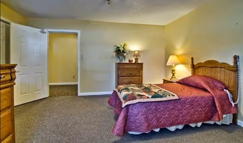 A bedroom with a wooden bed frame and a maroon quilted bedspread. There is a wooden dresser with a lamp and a flower arrangement on top. The room has beige walls and carpeted floor. An open door leads to another room with similar beige walls.