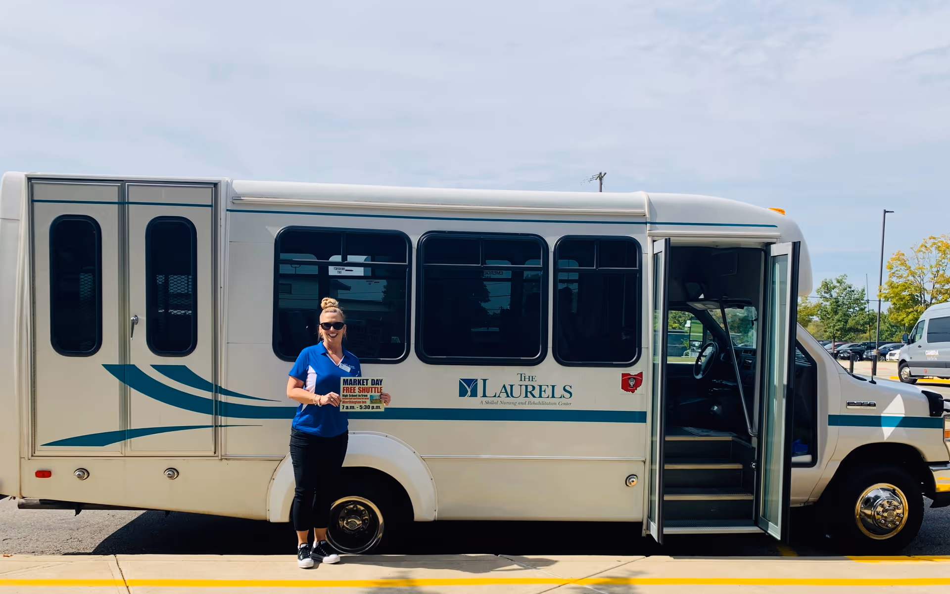 A woman wearing sunglasses and a blue shirt stands in front of a white shuttle bus with teal stripes and the logo 'The Laurels' on its side. The shuttle door is open, and the woman is holding a sign that reads 'Market Day Free Shuttle 7 a.m. - 5:30 p.m.'. The scene is outdoors with a parking lot and trees in the background.