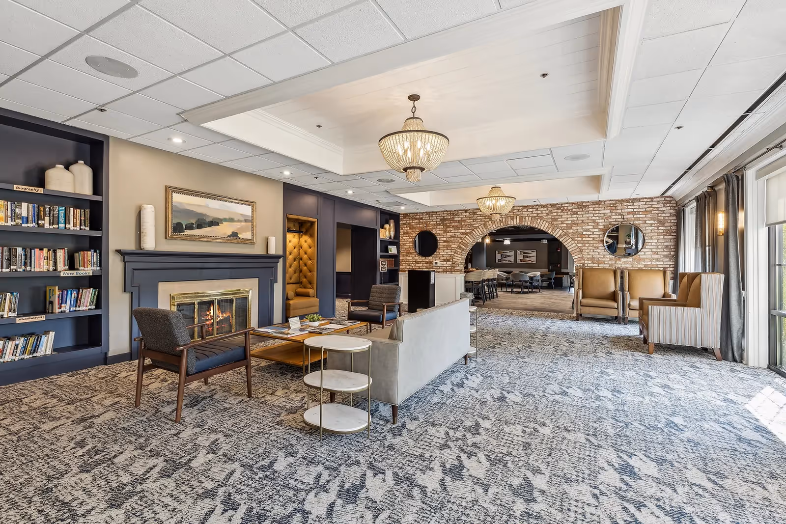 A spacious and well-lit living room area in The Lafayette featuring a patterned carpet, a fireplace with a painting above it, built-in bookshelves, comfortable seating including armchairs and a sofa, two chandeliers, and a brick archway leading to another room with additional seating and tables. Large windows with curtains allow natural light to fill the space.