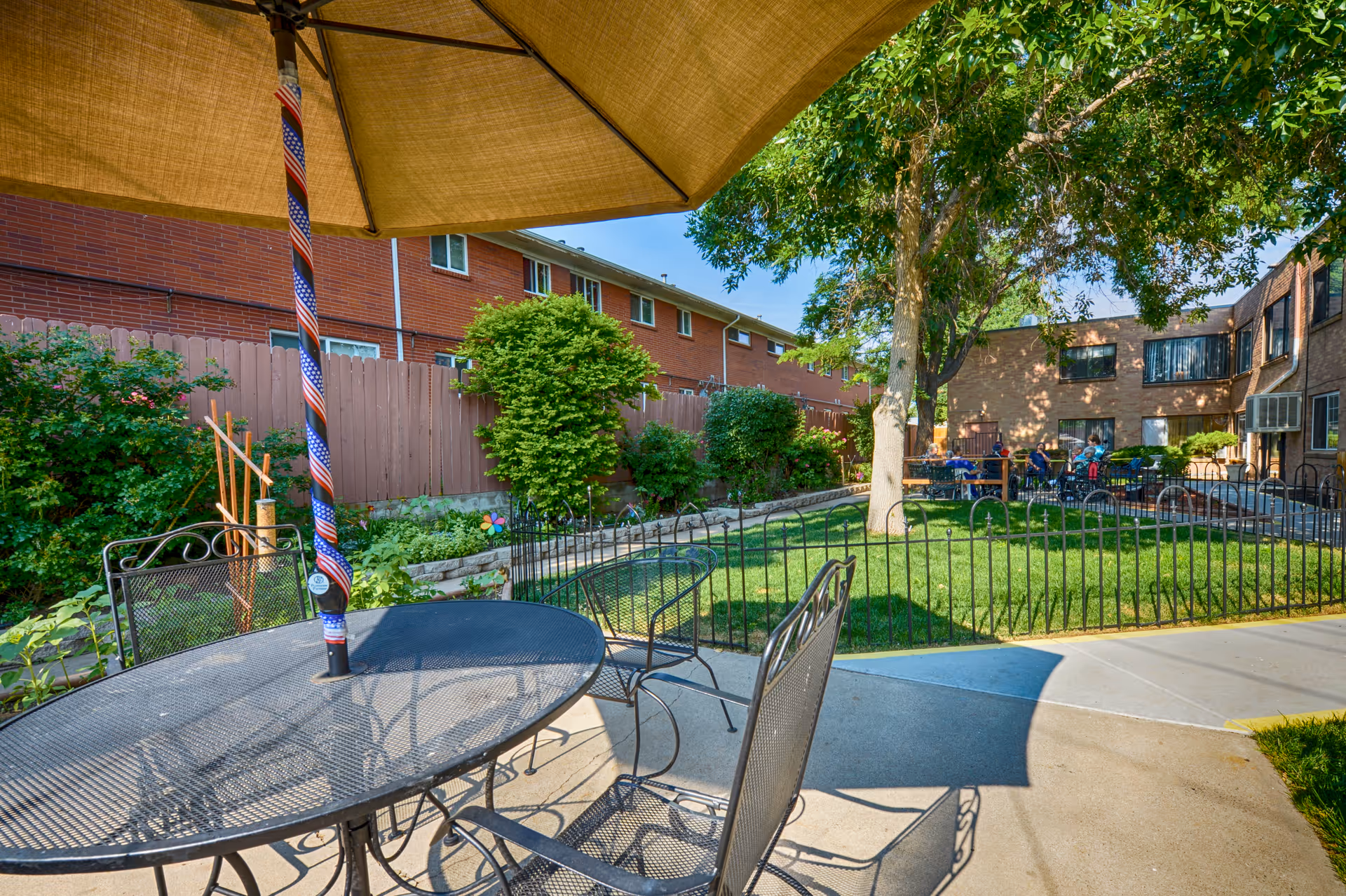Outdoor courtyard with a metal patio table and umbrella overlooking a fenced grassy garden and surrounding brick buildings.