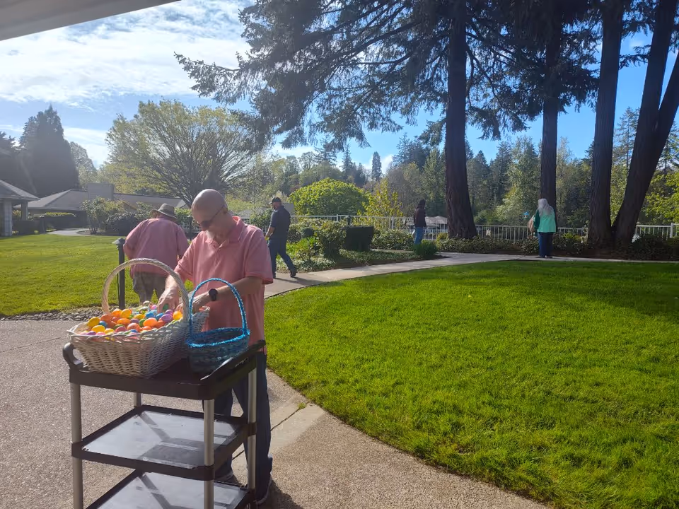 A man in a pink shirt standing outdoors next to a cart with baskets filled with colorful plastic eggs. In the background, several people are walking or standing on a paved pathway surrounded by green grass, trees, and bushes under a partly cloudy sky.