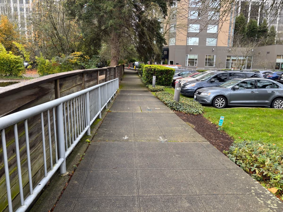 Sidewalk lined with a metal railing and landscaping, with parked cars and a multi-story building to the right.