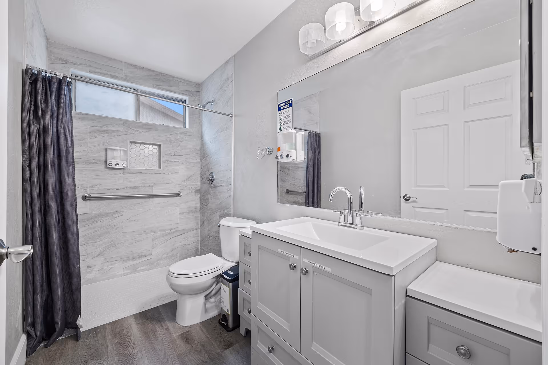 A clean and modern bathroom with a walk-in shower featuring gray tiles and a dark shower curtain. There is a white toilet next to a gray vanity with a white sink and a large mirror above it. The bathroom has wood-style flooring and a white door reflected in the mirror.