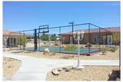 Outdoor basketball court with a fenced perimeter located between single-story buildings with red-tiled roofs. There is a paved walkway and a white lamp post in the foreground, surrounded by desert landscaping with rocks and sparse vegetation.