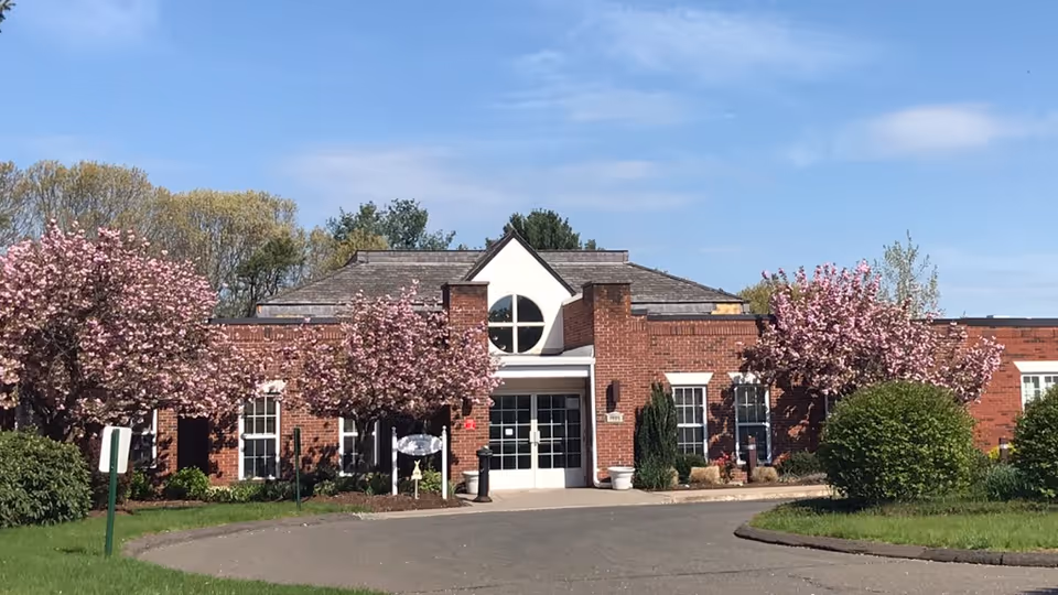Front exterior view of a single-story brick building with a peaked roof and a large circular window above the entrance. The entrance has double glass doors and is flanked by blooming pink cherry blossom trees and green shrubs. The sky is clear and blue.