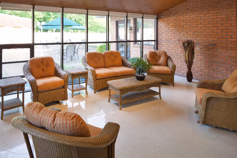 A sunlit sitting area with wicker furniture including two armchairs, a loveseat, a sofa, and a coffee table with a potted plant. The room has large windows showing an outdoor patio with tables and umbrellas, and a brick wall on one side.
