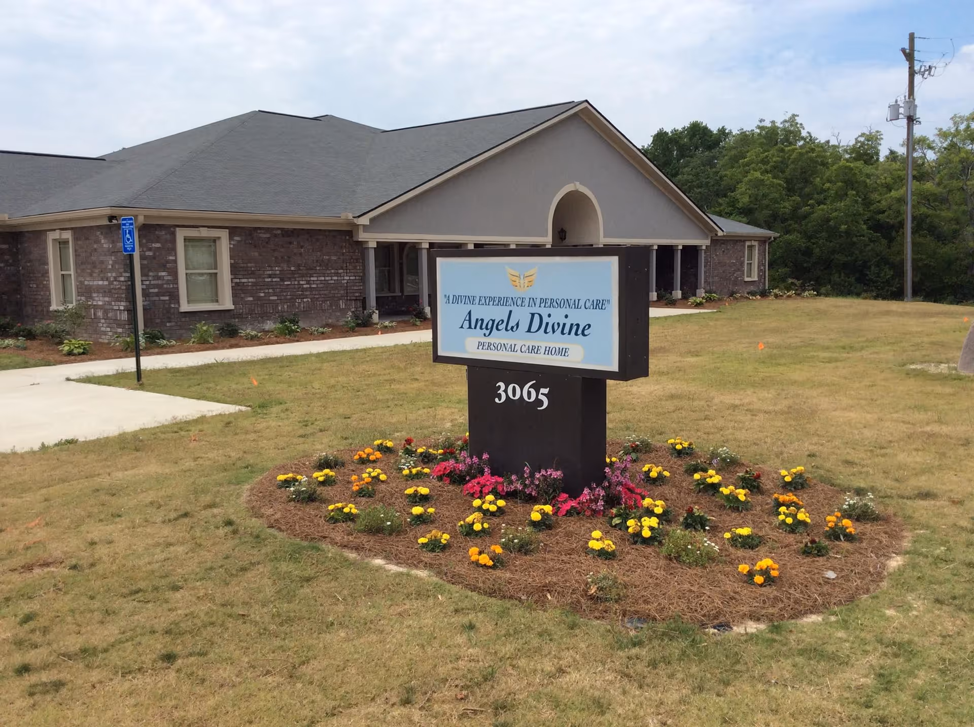Exterior view of Angels Divine Personal Care Home building with a sign in front displaying the facility name and address 3065, surrounded by a flower bed with yellow and pink flowers.