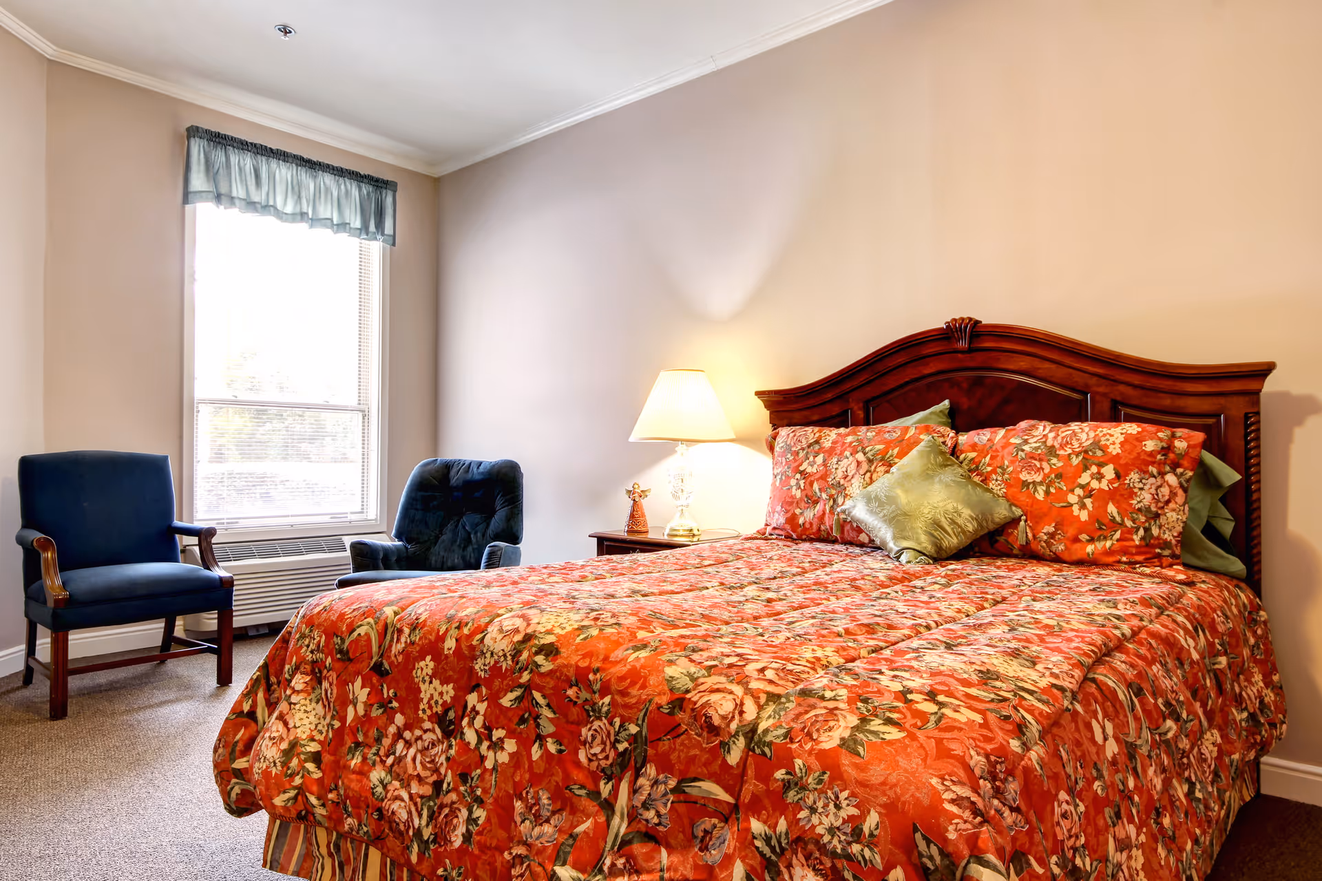 A cozy bedroom with a wooden headboard, floral-patterned bedspread, a bedside table with lamp, and two chairs by a window.