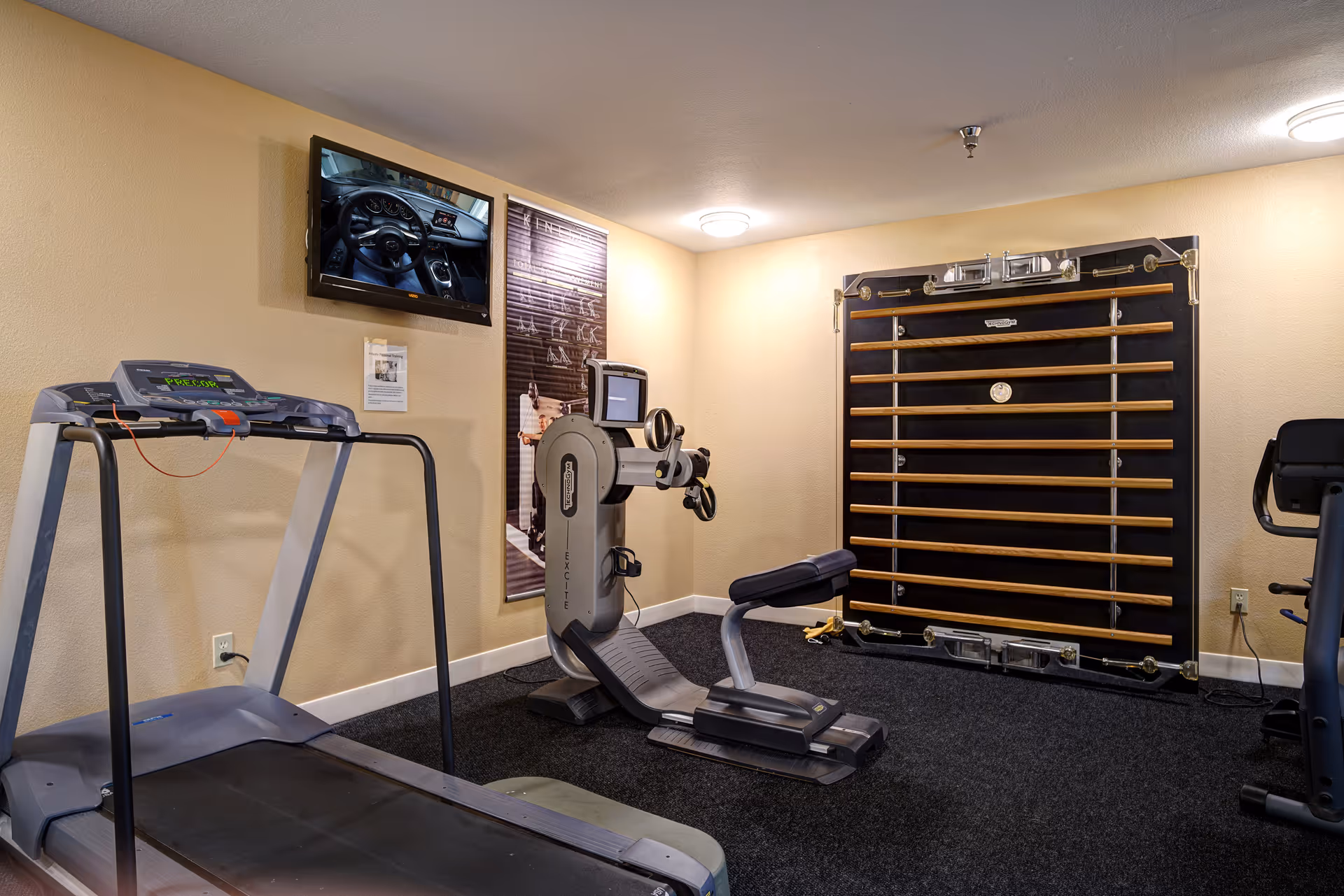 A small fitness room with exercise equipment including a treadmill, a seated exercise machine, and a stationary bike. There is a wall-mounted TV displaying a car dashboard, a poster on the wall, and a wooden wall-mounted exercise apparatus. The room has beige walls and dark carpeted flooring.