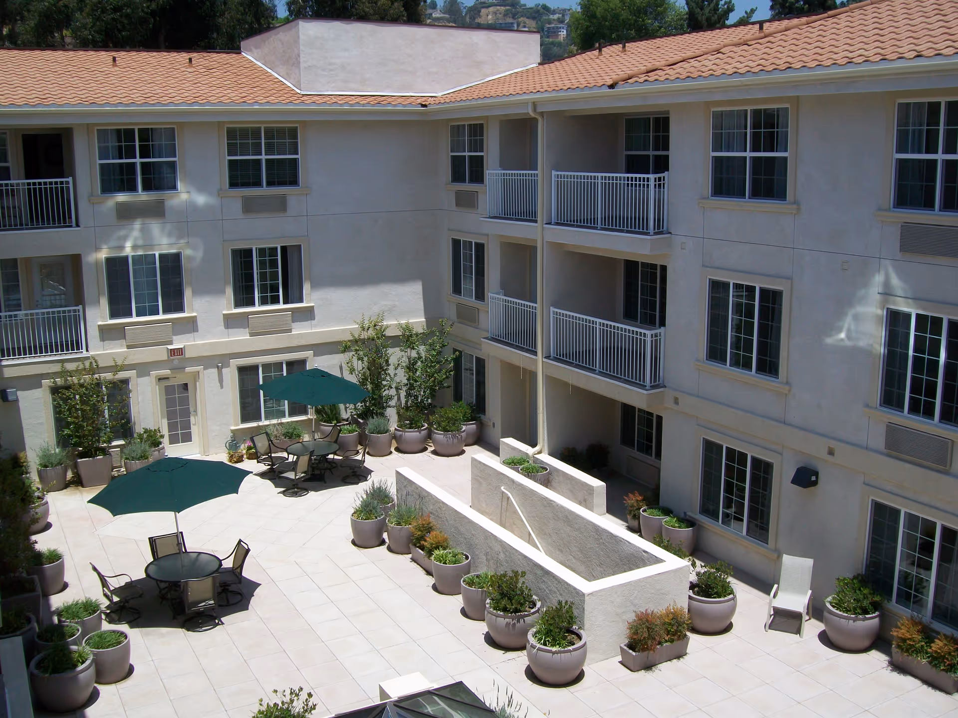 Outdoor courtyard area of a senior living facility with multiple potted plants, patio tables with green umbrellas, and chairs. The courtyard is surrounded by a three-story building with balconies and windows.