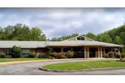 Exterior view of a single-story healthcare facility building with a covered entrance, surrounded by greenery and trees under a cloudy sky.