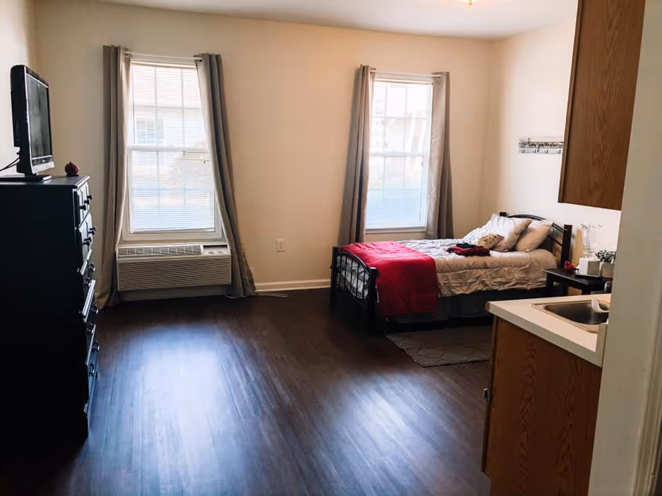 A small, simple bedroom with two large windows covered by beige curtains. The room has dark wooden flooring, a single bed with white bedding and a red blanket, a small nightstand with a lamp and some decor, a black dresser with a TV on top, and a small kitchenette area with a sink and wooden cabinets.