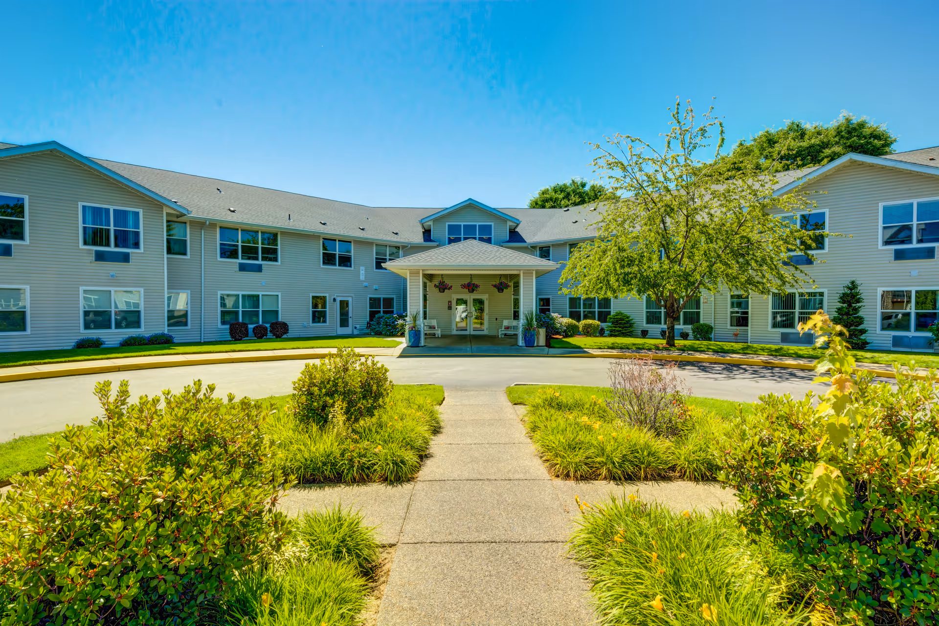 Front exterior view of The Suites Assisted Living and Memory Care building with a covered entrance, surrounded by green shrubs, trees, and a clear blue sky.