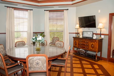Bright dining room with a round wooden table surrounded by upholstered chairs, a floral centerpiece, windows with curtains, and a sideboard with a wall-mounted TV.