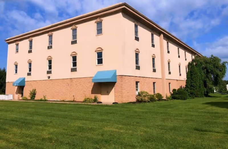 Exterior view of a three-story building with beige and light brown walls, multiple windows, and two blue awnings over doors. The building is surrounded by a well-maintained green lawn and some bushes, under a partly cloudy blue sky.