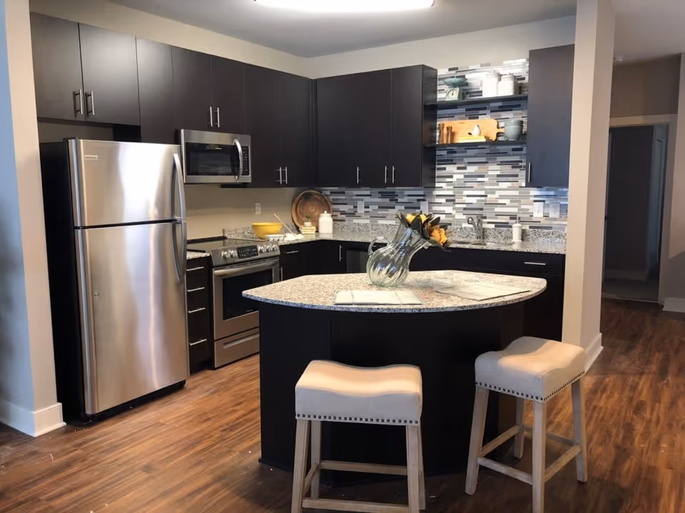 Modern kitchen with dark cabinets, stainless steel refrigerator, oven, and microwave. There is a curved granite countertop island with two beige cushioned stools and a glass vase with flowers on top. The backsplash features a mosaic tile design in shades of gray, white, and black. The floor is wood, and there is an open doorway leading to another room.
