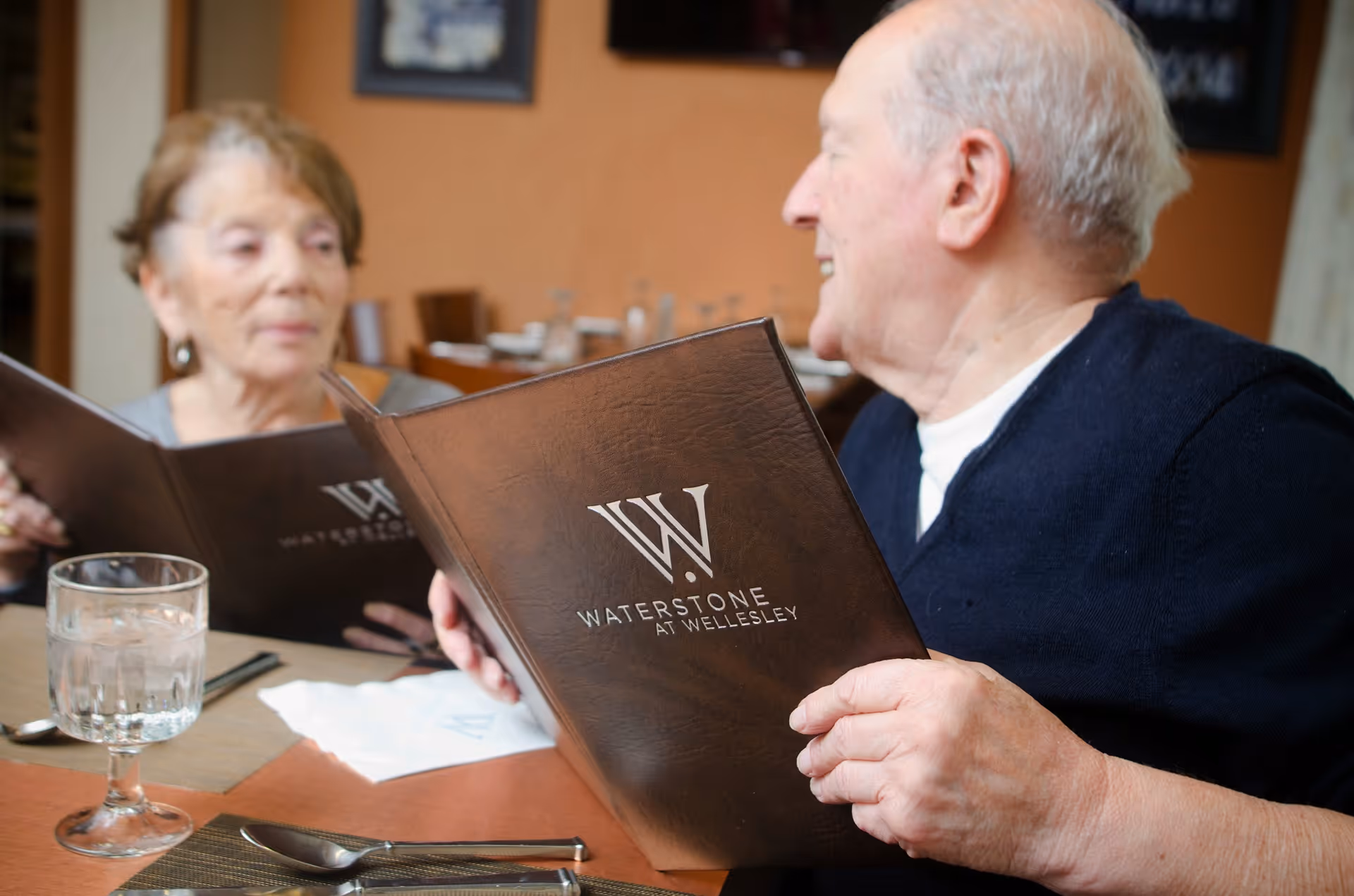 An elderly man and woman sitting at a dining table in a restaurant or dining area, each holding a menu from Waterstone At Wellesley. The table is set with a glass of water, utensils, and a napkin.