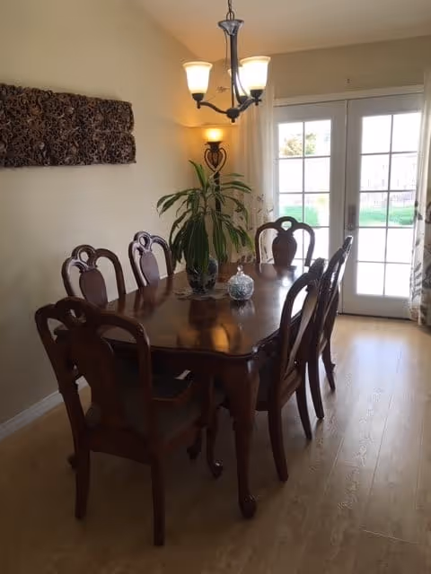 Wooden dining table with six chairs, a potted plant centerpiece, chandelier lighting, and French doors letting in natural light.