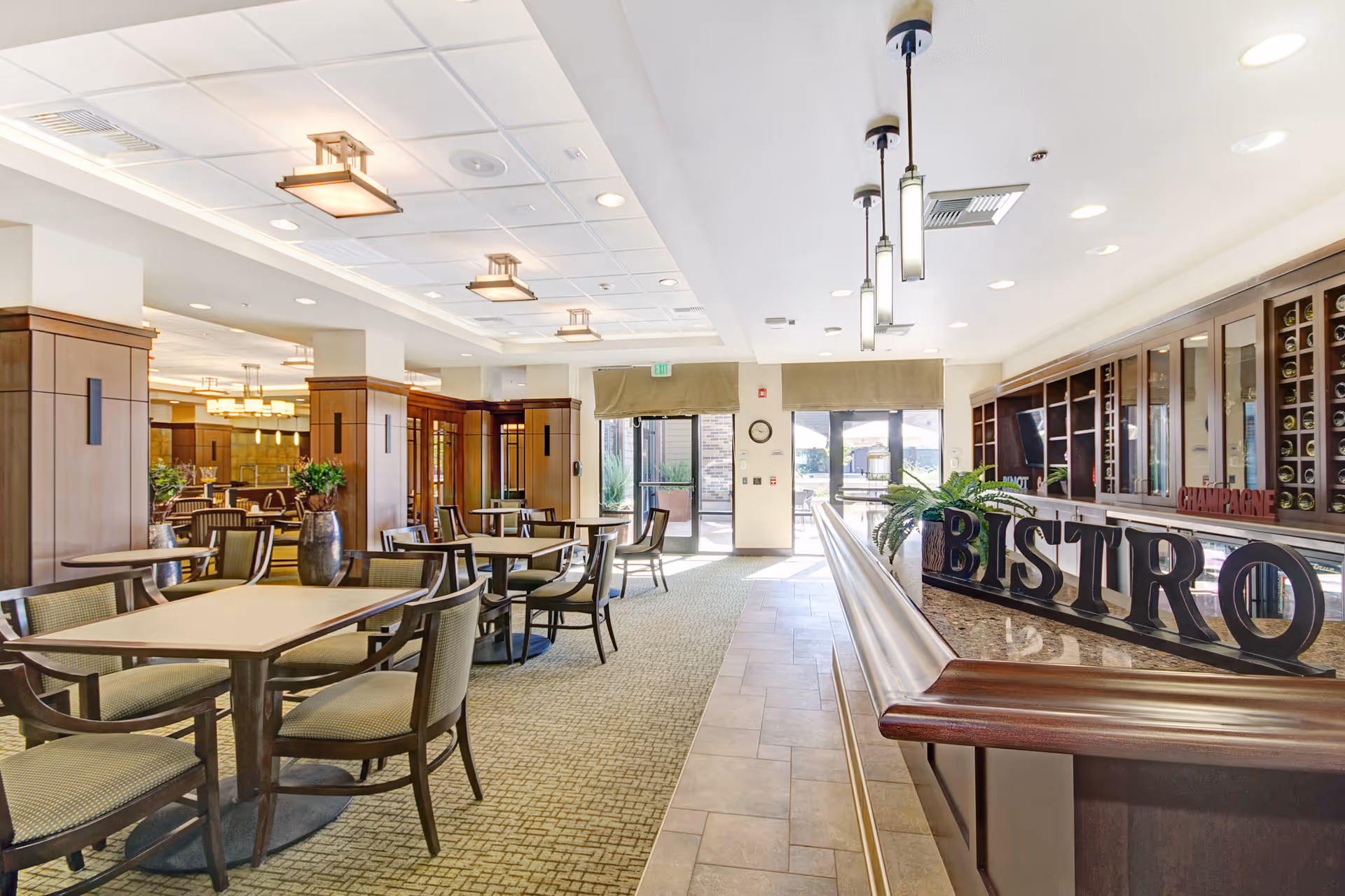 Bright and spacious dining area with multiple tables and chairs, wooden paneling, and modern light fixtures. A counter with a decorative 'BISTRO' sign and plants is visible on the right side, with large windows and glass doors letting in natural light at the far end.