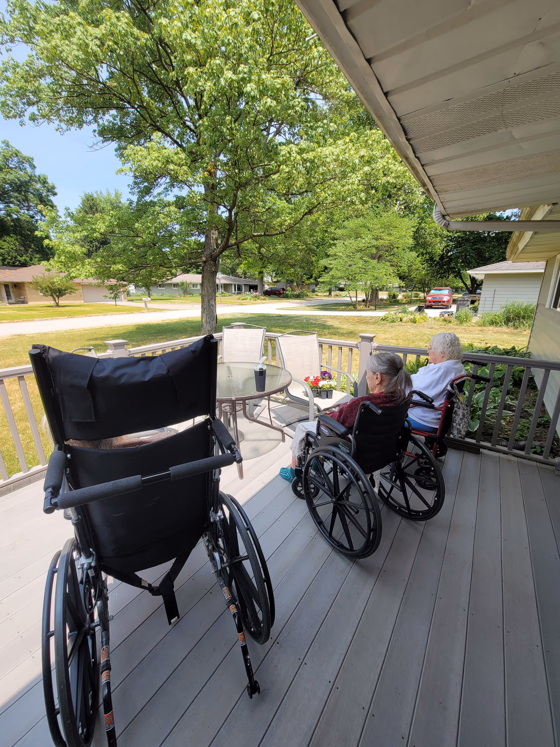 Two elderly women in wheelchairs sitting on a wooden porch with a glass table and chairs, overlooking a green yard with trees and a quiet neighborhood street.