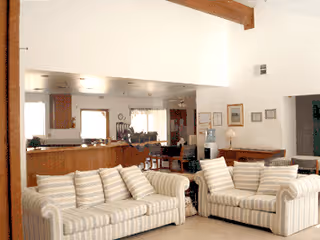 A spacious living room with two striped sofas facing each other, a wooden coffee table between them, and a high ceiling with exposed wooden beams. In the background, there is a kitchen area with wooden cabinets and a dining table with chairs. The room is well-lit with natural light coming through windows.