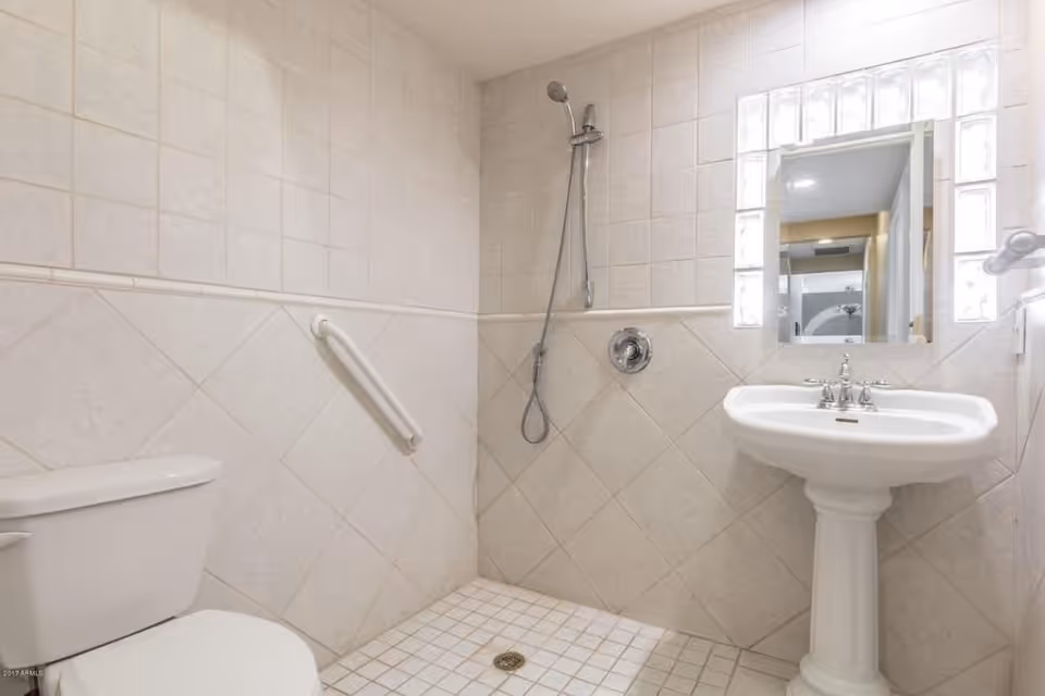 A clean, tiled bathroom with a white toilet, a pedestal sink with a mirror above it, and a walk-in shower area with a handheld showerhead and a grab bar on the wall.