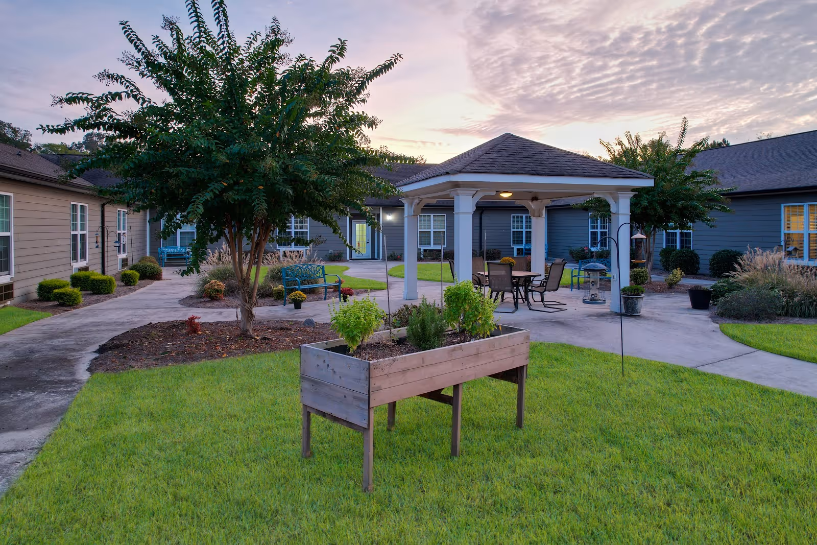 A landscaped outdoor courtyard with a gazebo, patio seating, a raised planter box, and surrounding single-story residence buildings at sunset.