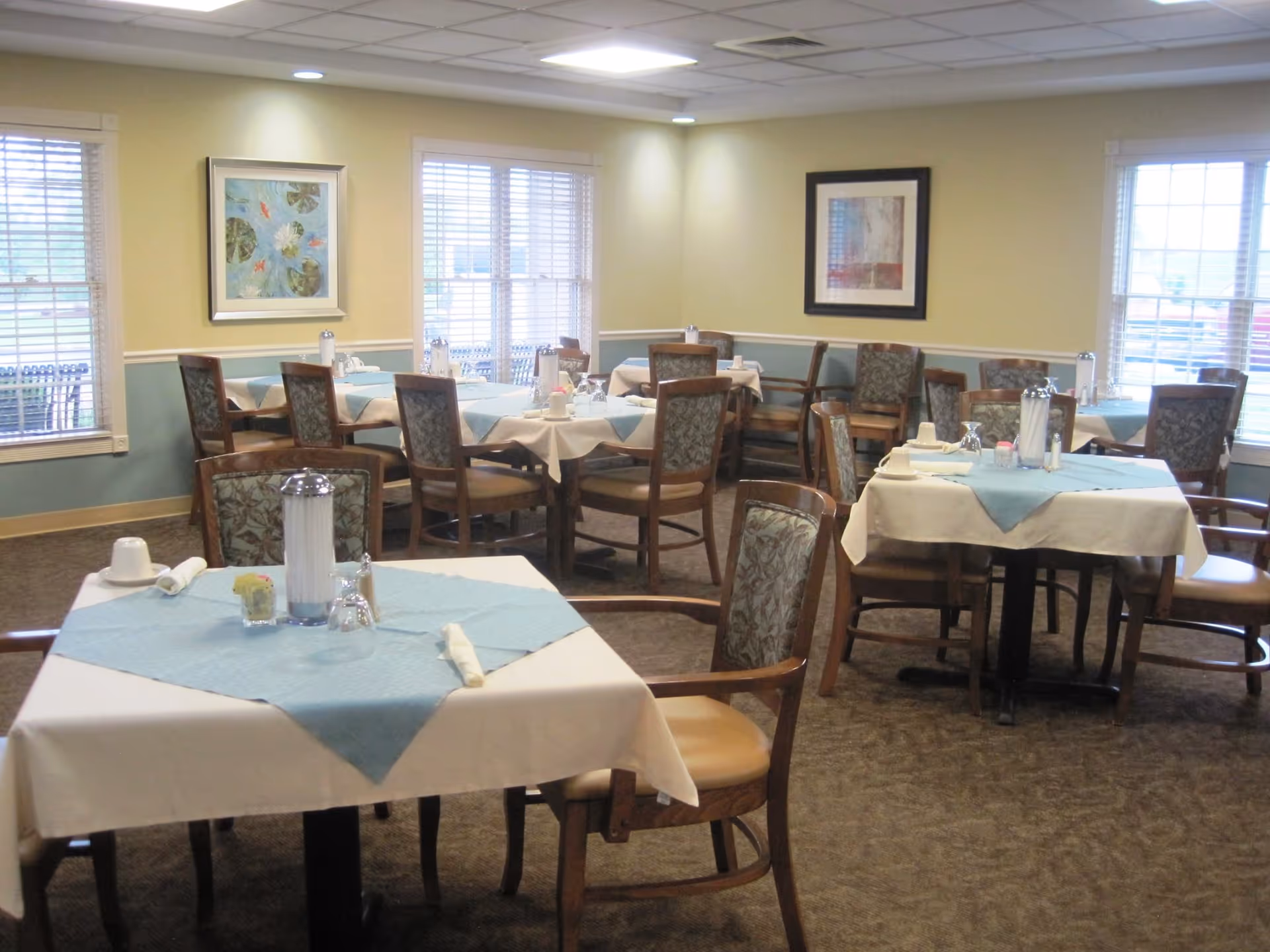 Dining room with multiple tables set with white tablecloths and blue napkins, wooden chairs, and framed artwork on the walls.