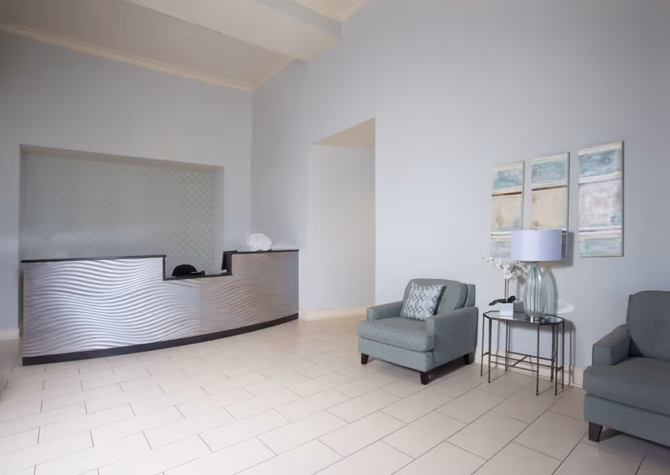 A modern reception area with a wavy textured front desk, two upholstered armchairs, a small side table with a glass lamp and a white orchid, and three abstract paintings on a light blue wall.