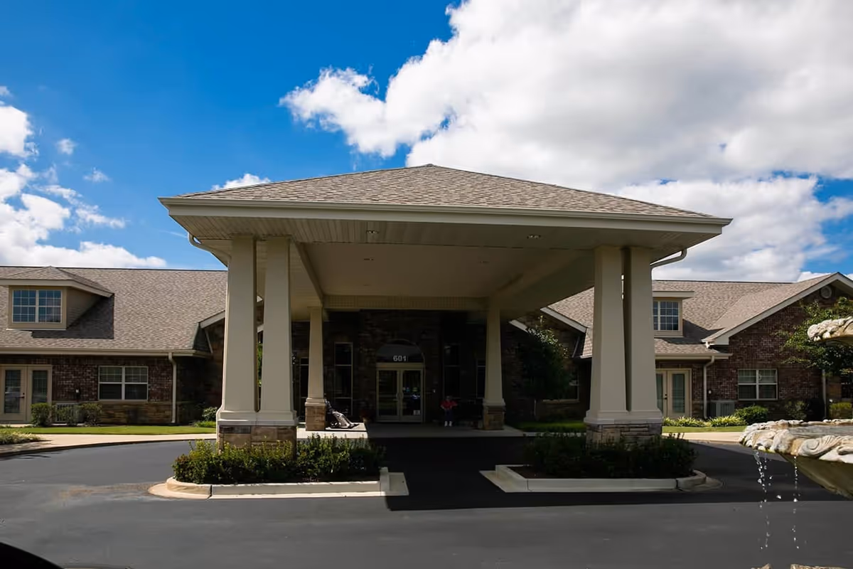 Front entrance of a senior living facility with a covered driveway supported by four large columns. The building has a brick exterior with multiple windows and a gray shingled roof. A water fountain is partially visible on the right side of the image under a partly cloudy blue sky.
