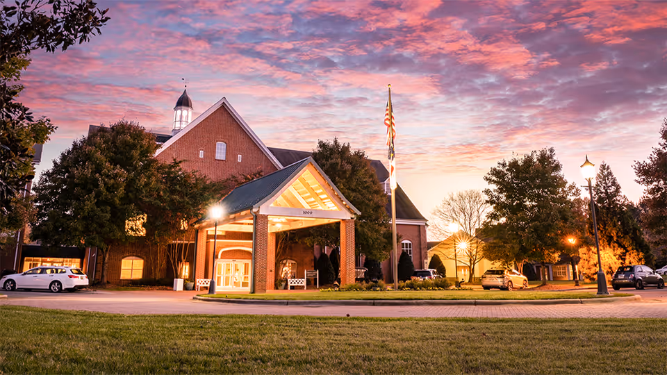Exterior view of Salemtowne Retirement Community building at dusk with a colorful sky, a covered entrance with lights, an American flag on a flagpole, several trees, parked cars, and a well-maintained lawn.