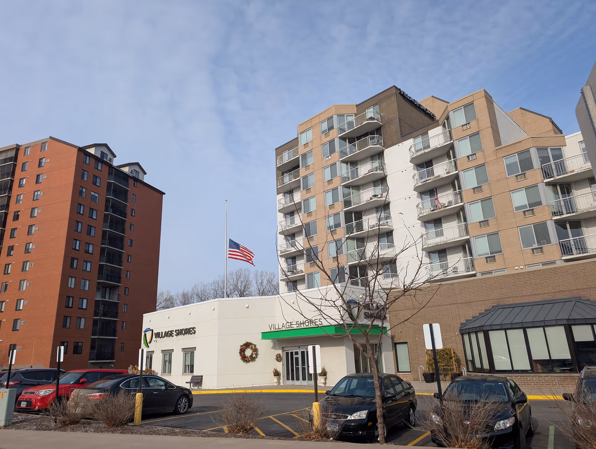 Exterior view of Village Shores senior living facility showing a multi-story building with balconies, an entrance with a green awning, an American flag at half-mast, several parked cars, and a clear sky.
