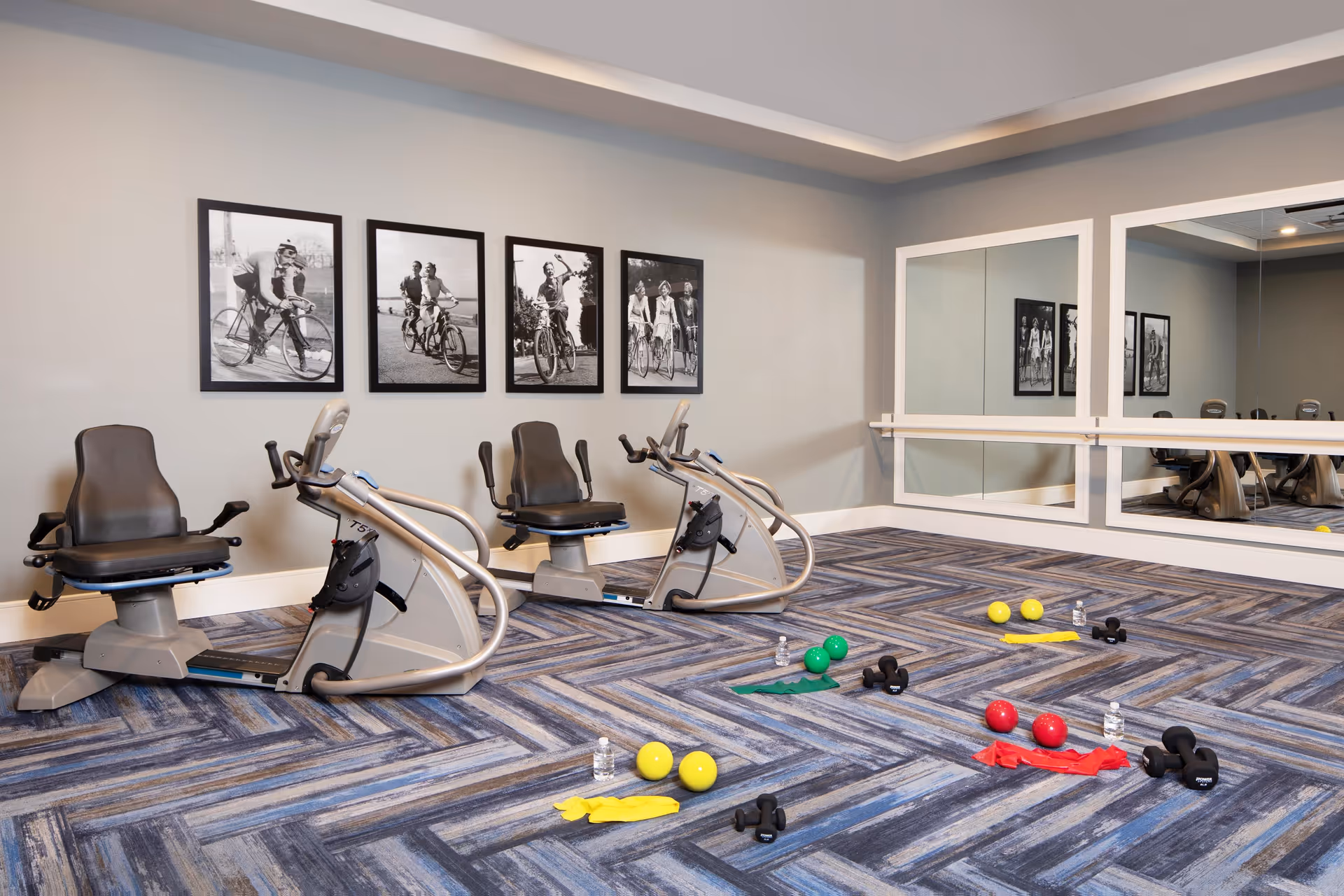 Bright exercise room with recumbent bikes, large mirrors, framed cycling photos, and small weights and exercise equipment on patterned carpet.