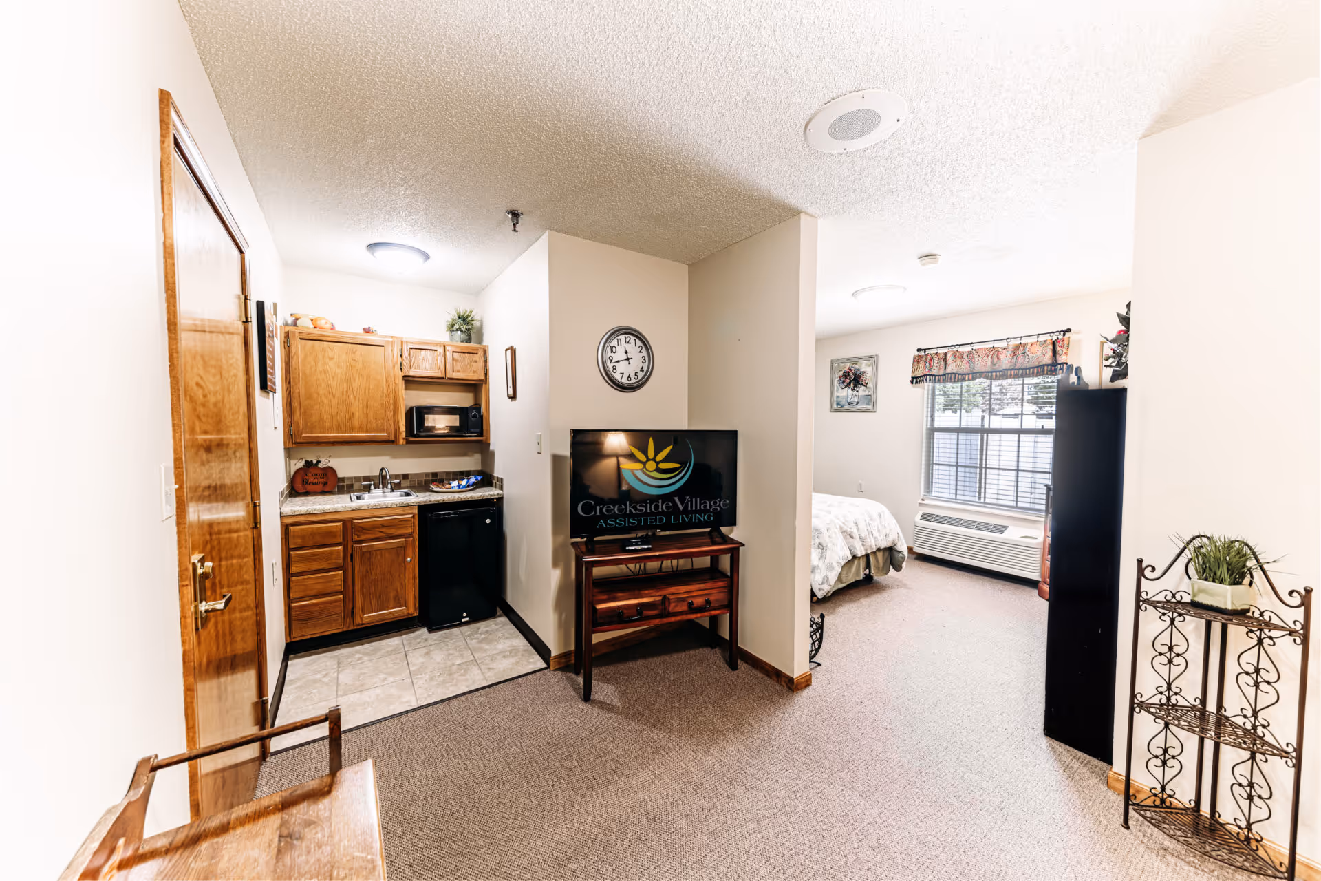 Interior view of a senior living facility apartment at Creekside Village featuring a small kitchenette with wooden cabinets, a microwave, a sink, and a mini refrigerator. Adjacent to the kitchenette is a wall-mounted clock and a TV on a wooden stand displaying the Creekside Village Assisted Living logo. In the background, there is a bedroom area with a bed, a window with a decorative valance, an air conditioning unit below the window, and some wall art. The room has carpeted flooring and neutral-colored walls.