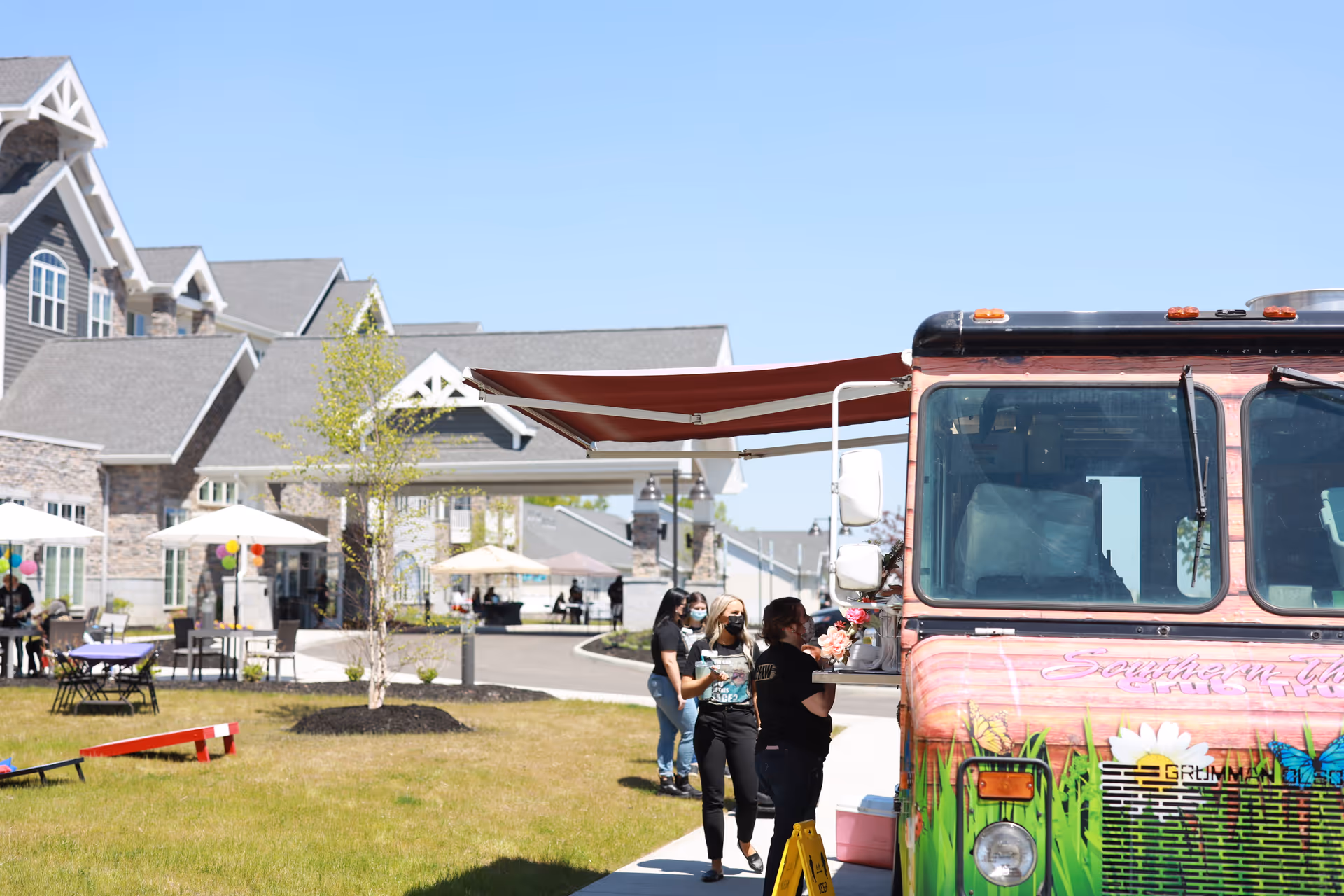 A colorful food truck at a senior living community courtyard with people in line, outdoor tables and umbrellas, and the community building in the background.