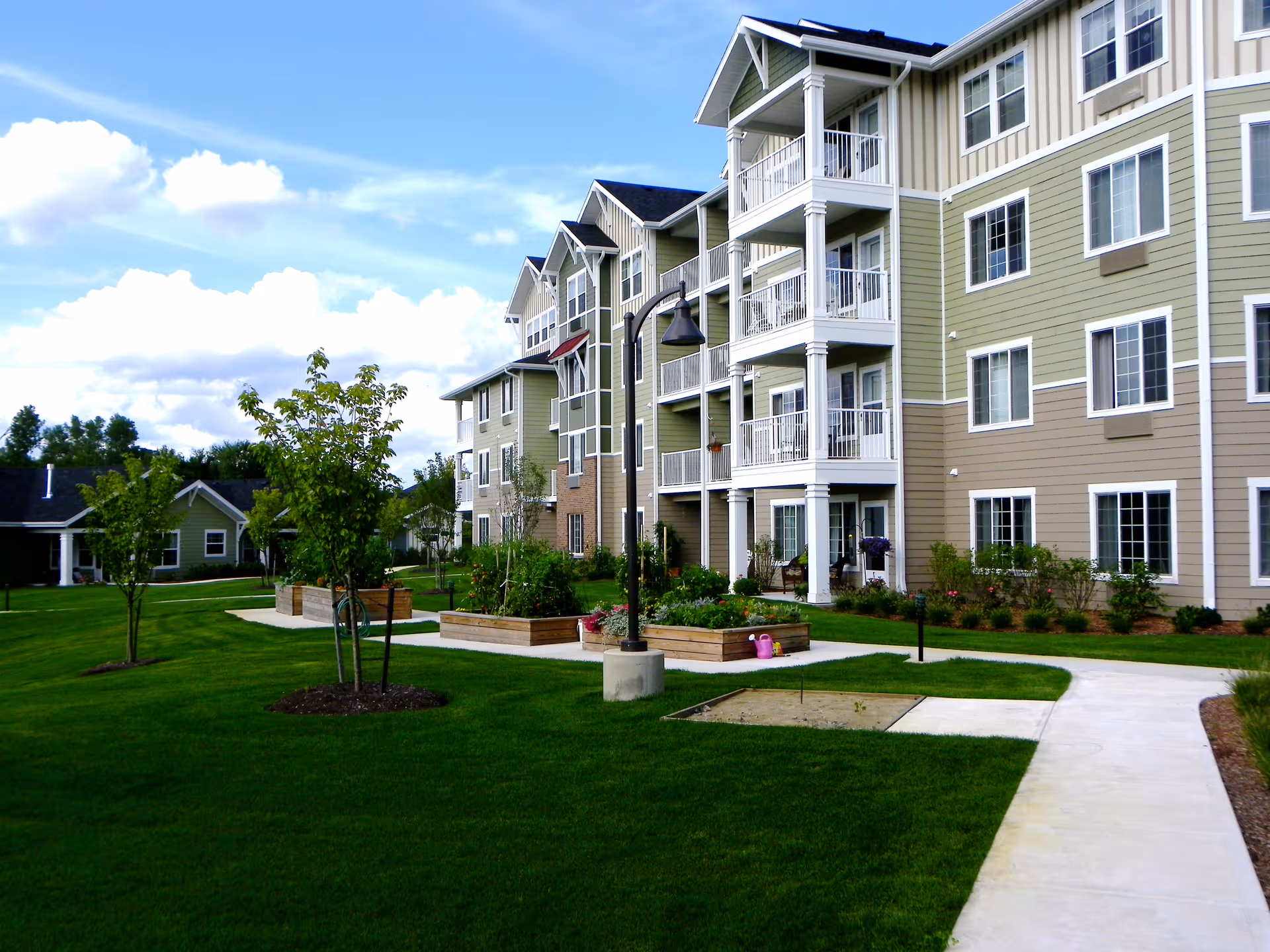 Exterior view of a multi-story green and beige senior living building with balconies, landscaped lawn, walkways, and raised garden beds.
