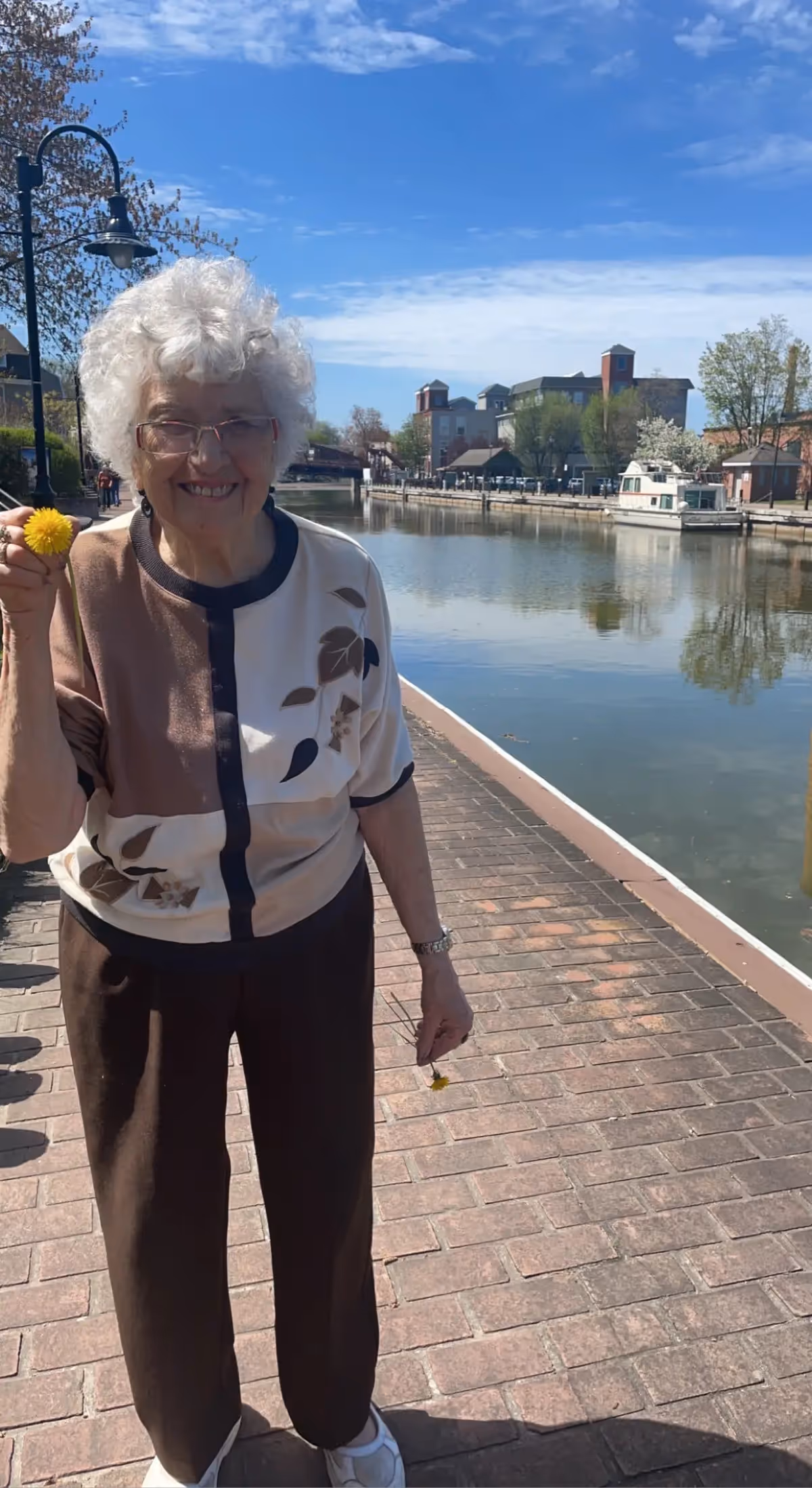 An elderly woman with white curly hair and glasses stands on a brick pathway beside a calm river or canal. She is smiling and holding a yellow flower in her right hand. The background shows buildings, trees, and a boat on the water under a blue sky with some clouds.