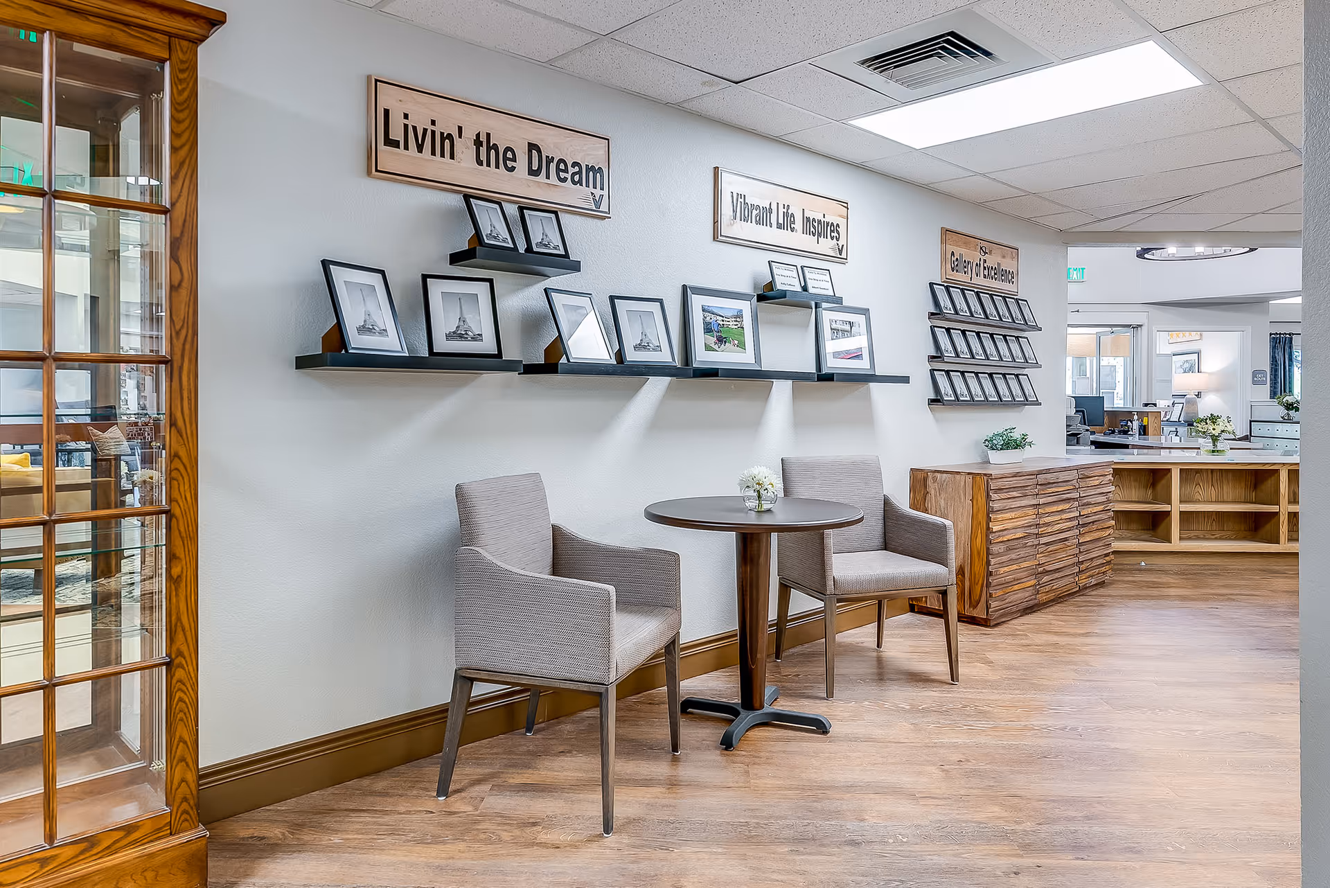 Small seating area in a senior living facility lobby with two chairs around a round table, framed photos on wall shelves, and decorative wooden cabinets.