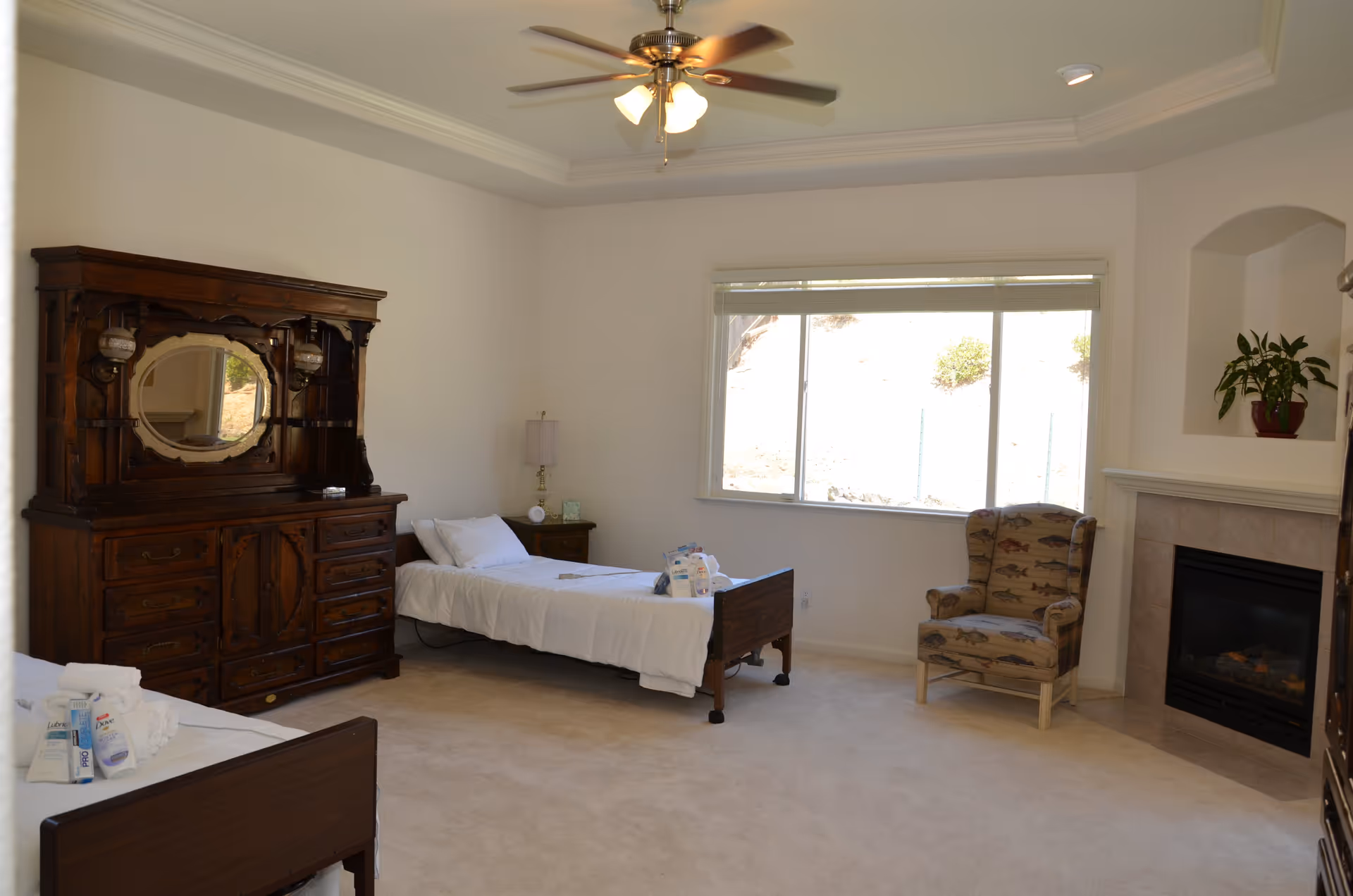 A spacious bedroom with two single beds, each with white bedding and toiletries placed on top. There is a large wooden dresser with an ornate mirror on the left side, a nightstand with a lamp between the beds, a patterned armchair near a fireplace on the right, and a large window letting in natural light. The room has beige carpet and white walls with a ceiling fan overhead.
