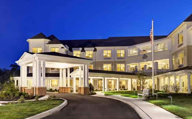 Exterior view of a large, well-lit senior living facility building at dusk with a covered entrance, multiple windows, an American flag, and landscaped grounds.