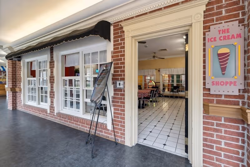 Entrance to The Ice Cream Shoppe with a brick exterior wall, white framed windows, and a black awning. A sign with an ice cream cone graphic and the words 'The Ice Cream Shoppe' is mounted on the wall next to the open door, revealing a tiled floor and tables with checkered tablecloths inside.