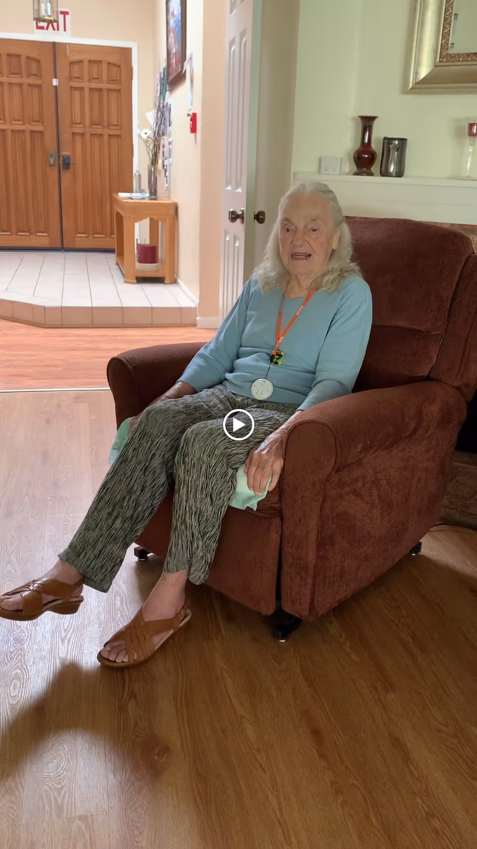 An elderly woman with white hair is sitting in a brown armchair in a room with wooden flooring. She is wearing a light blue long-sleeve shirt, patterned pants, and brown sandals. Behind her is a white wall with decorative vases on a shelf and a framed picture. In the background, there is a hallway with double wooden doors and a small wooden table with items on it.